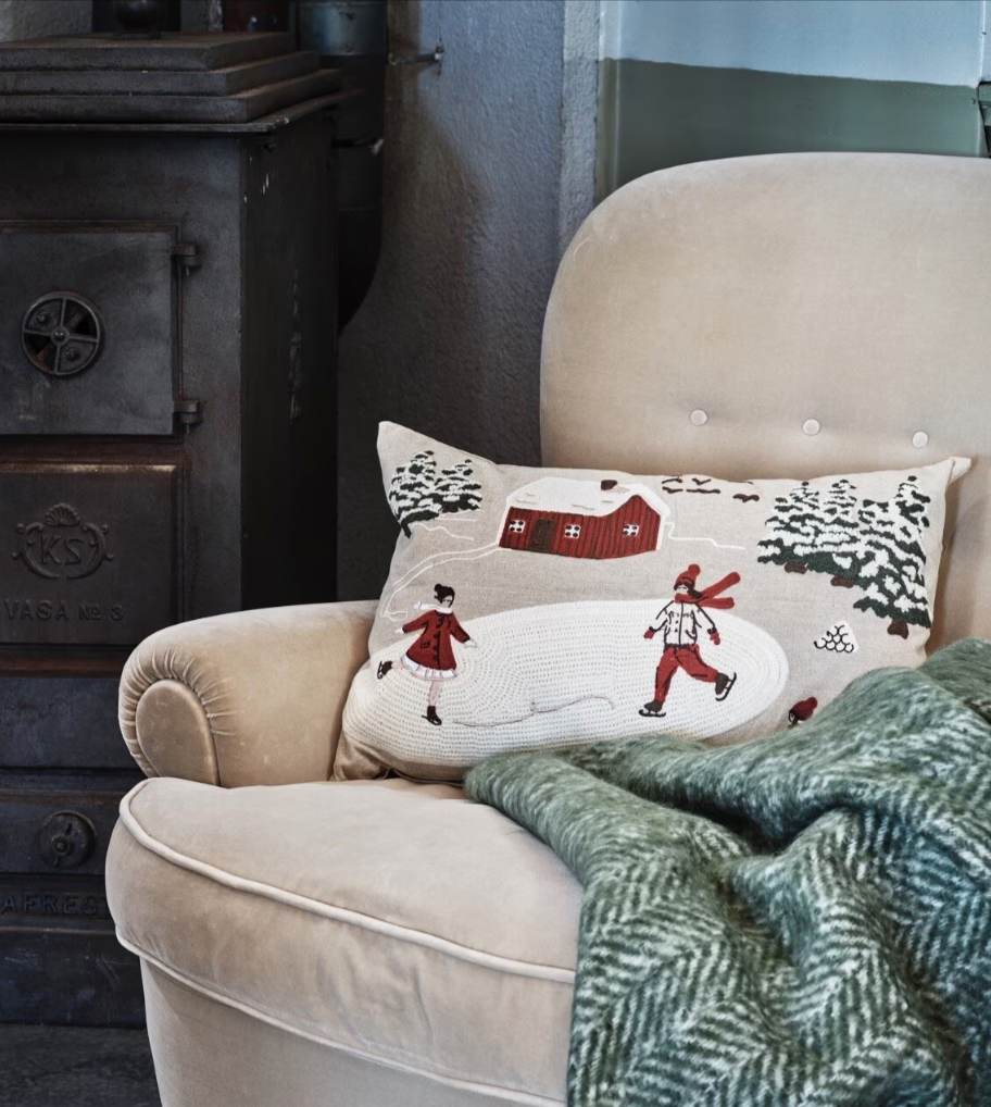 A beige armchair with a green patterned blanket and the Strömshaga Cushion Cover Vinterland Embroidered, 40 x 60 cm, sits next to a dark metal wood stove.
