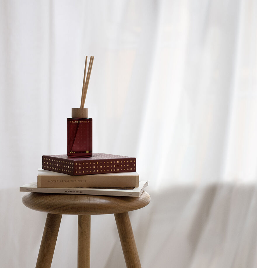 A Skandinavisk Jul Scent Diffuser (100 ml) and two hardcover books sit on a round wooden stool before sheer white curtains, setting a cozy Scandinavian Christmas atmosphere.