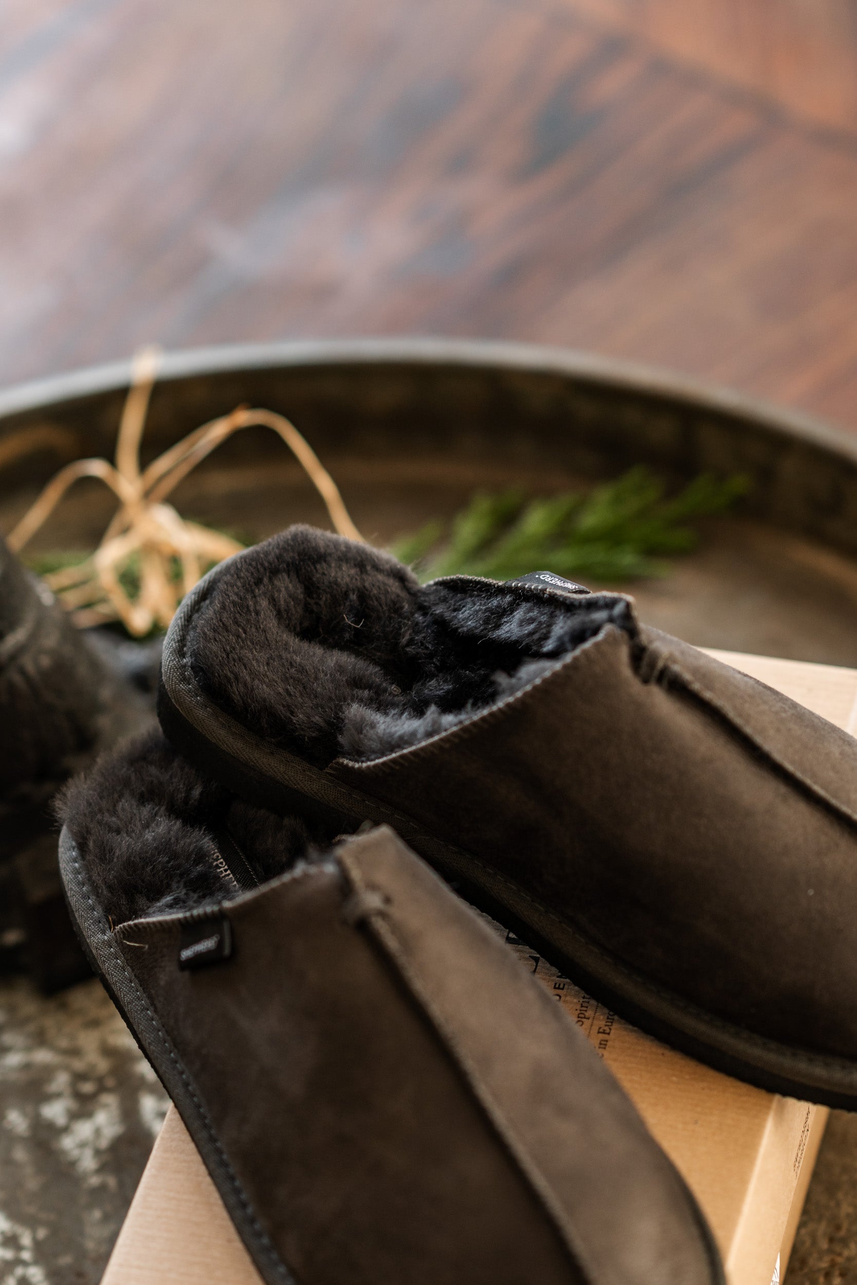 A pair of Shepherd of Sweden Hugo men's sheepskin slippers in Asphalt Grey with a cozy fuzzy lining sits on a cardboard box, accented by greenery and a rustic tray. The easy slip-in design is soft, warm, and perfect for indoor comfort.