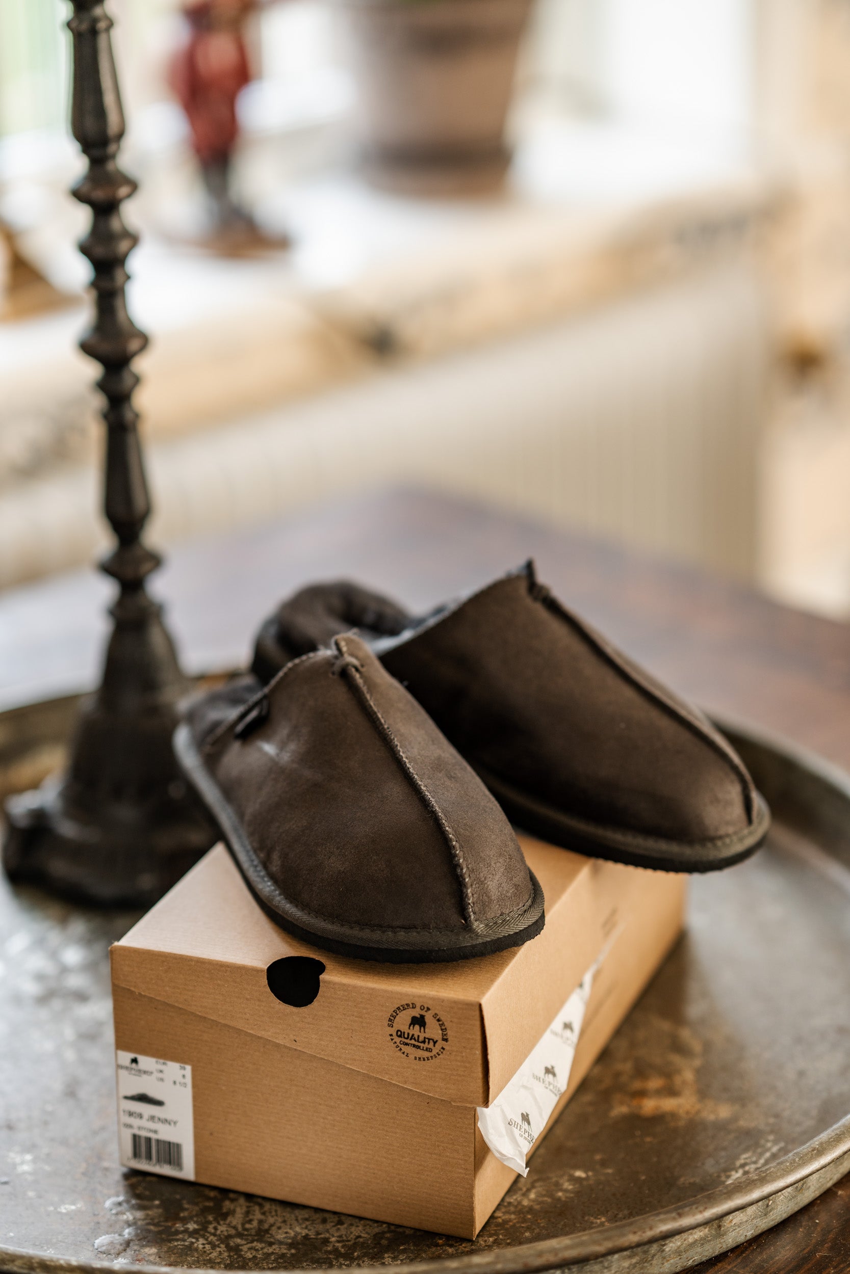 A pair of Shepherd of Sweden Hugo men's sheepskin slippers in Asphalt Grey sits atop a closed shoebox on a round metal tray placed on a wooden table, with a softly blurred background.