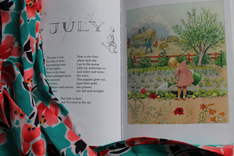 An open "Around the Year Mini Edition Book" by Elsa Beskow shows a July poem beside an illustration of children in a summer garden, with floral fabric partly covering this classic.