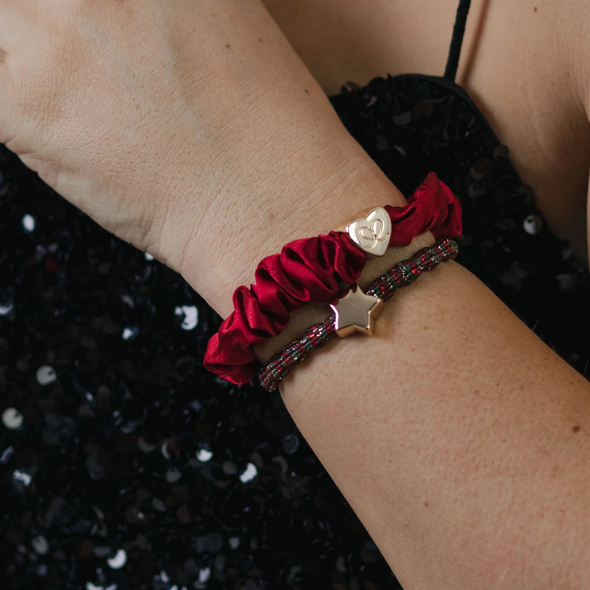 A person wears a ByEloise Silk Scrunchie in Burgundy Red with a gold heart along with a beaded bracelet featuring a star charm, paired with a black sequin top.