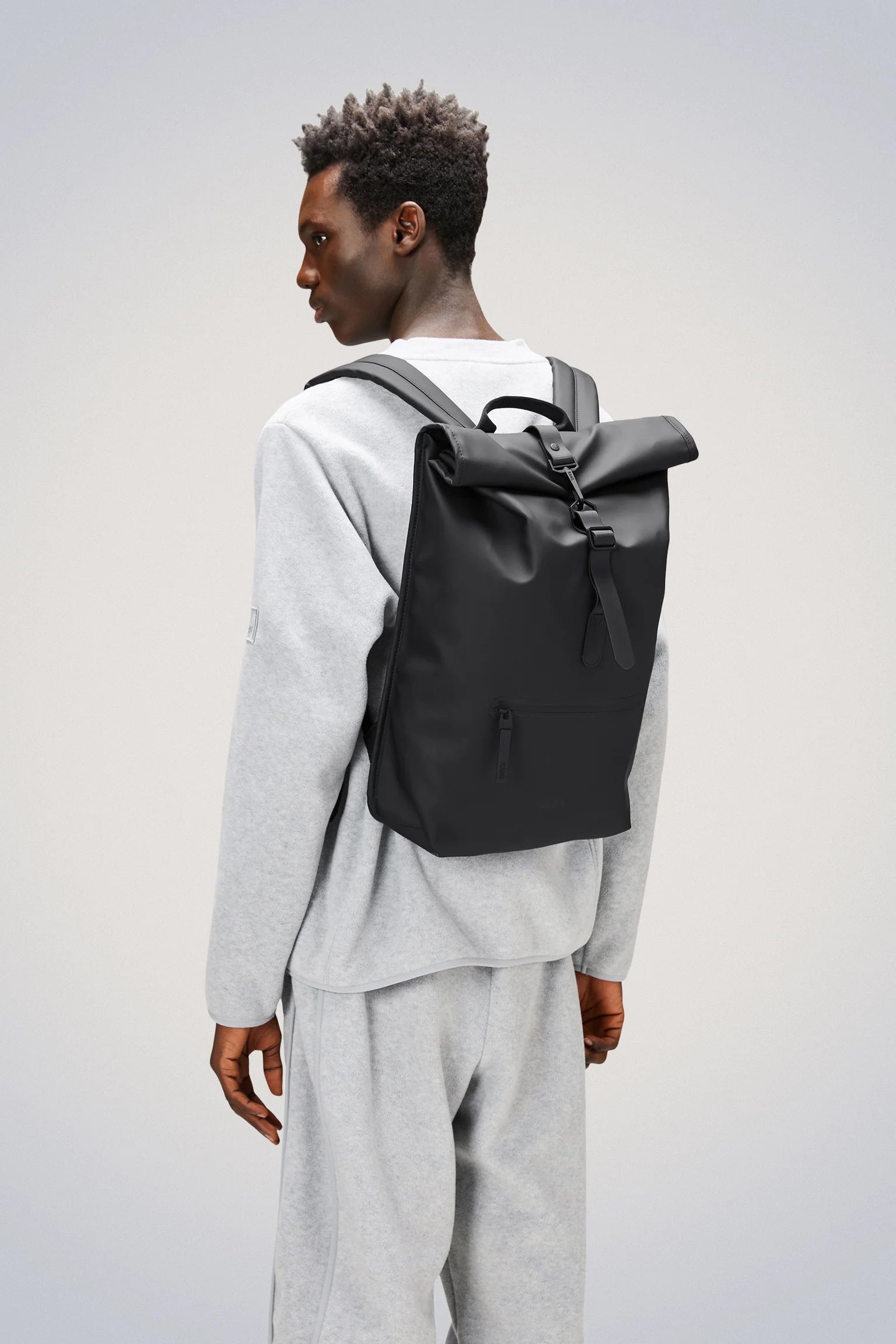 A person wearing a light gray tracksuit stands facing left against a plain background, carrying the Rains Rolltop Rucksack Backpack in Black by Rains.