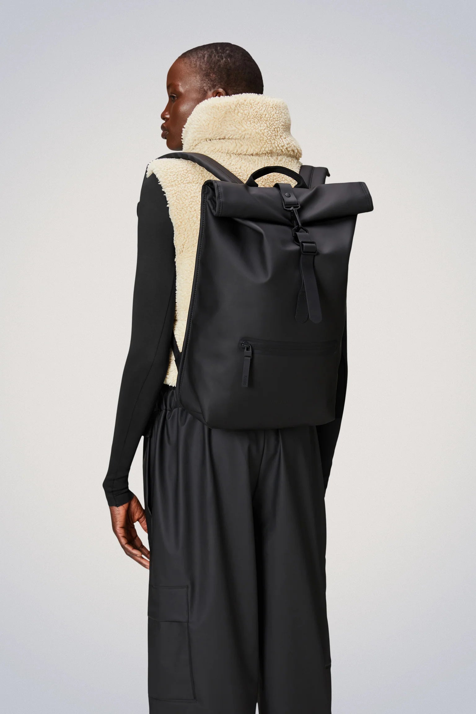 A person is shown from the back against a light gray background, wearing a cream fleece vest over a black long-sleeve shirt and black pants, carrying the Rains Rolltop Rucksack Backpack in Black by Rains, which features a large cycle-ready design and a front zipper pocket.