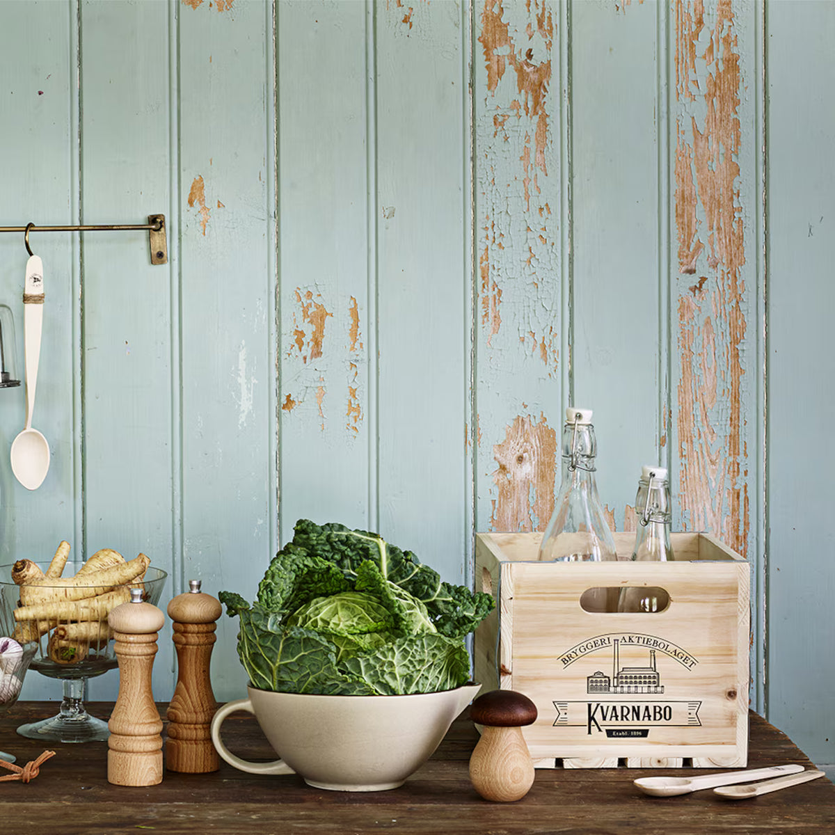 A rustic kitchen scene with a bowl of leafy cabbage, beech wood pepper and salt mills, root vegetables, the Strömshaga Nutcracker Mushroom in Brown Beech Wood, and a wooden crate holding glass bottles set against distressed blue wooden paneling.