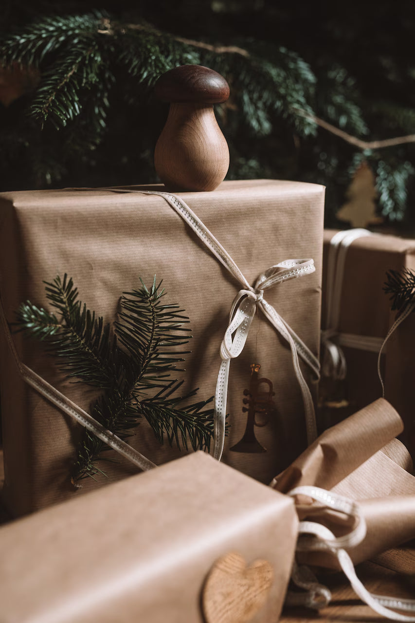 Gift boxes wrapped in brown paper and white ribbons, adorned with evergreen sprigs, are topped with the Strömshaga Nutcracker Mushroom in brown beech wood. A Christmas tree branch can be seen in the background.