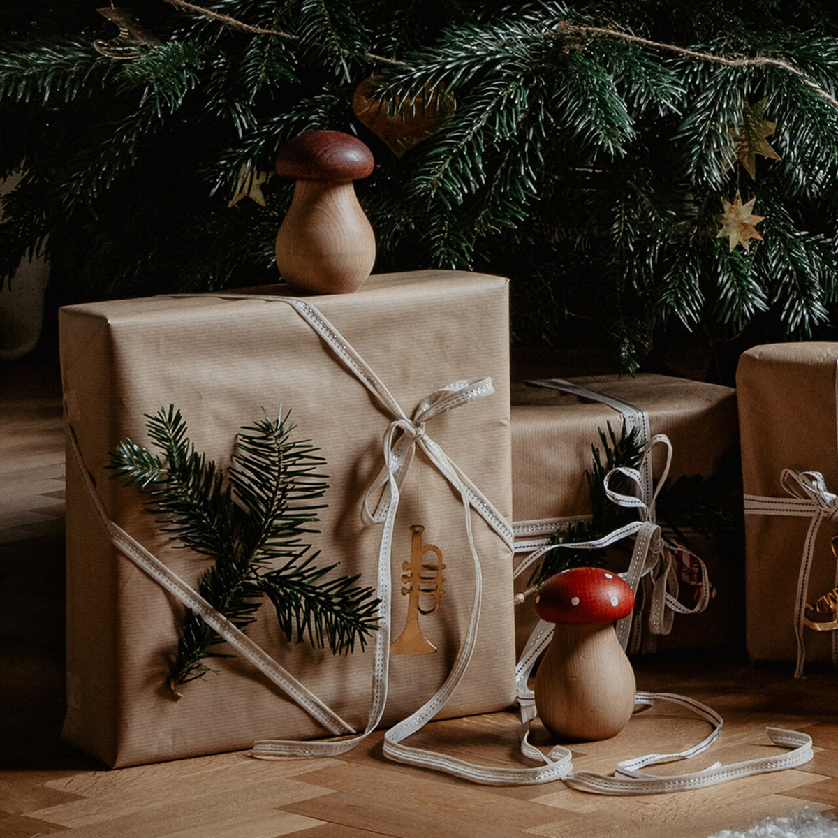 Gifts wrapped in brown paper with white ribbon and fir branches are arranged beneath a Christmas tree adorned with green needles and gold star ornaments, accompanied by charming Strömshaga Nutcracker Mushroom decorations in brown beech wood.