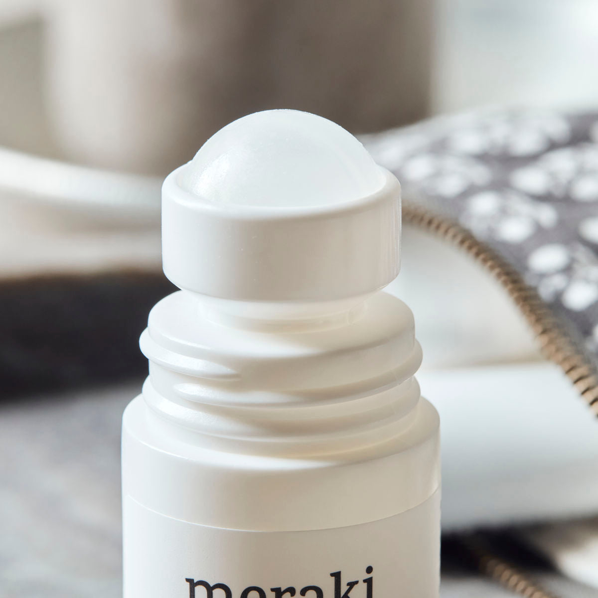 A close-up of a white Meraki Organic Deodorant, Linen Dew roll-on bottle, formulated with organic extracts, with the label partially visible next to a blurred patterned cloth in the background.