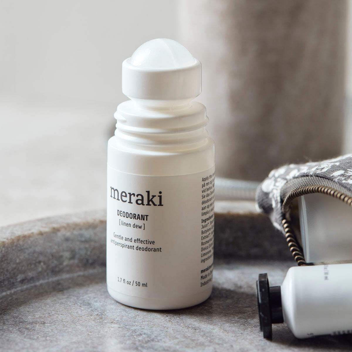 A white roll-on bottle of Meraki Organic Deodorant, Linen Dew sits on a round tray beside a patterned pouch and a small container. The focus is on the open deodorant, which features minimal, clean packaging and is formulated with organic extracts.