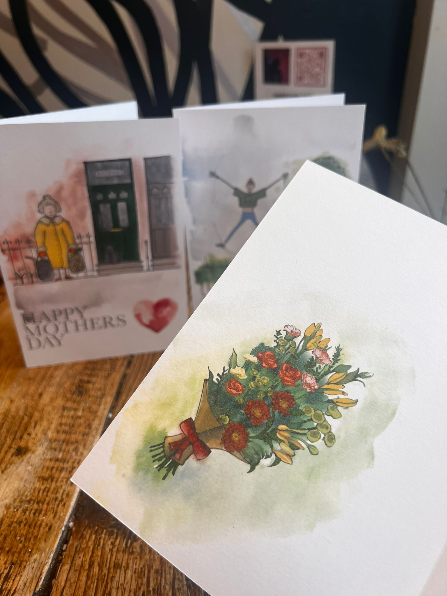 A close-up of the Blåbär Nordic Living “Mothers Day Card - Dog Mum,” featuring a watercolor flower bouquet, with other softly blurred cards in the background on a wooden surface.