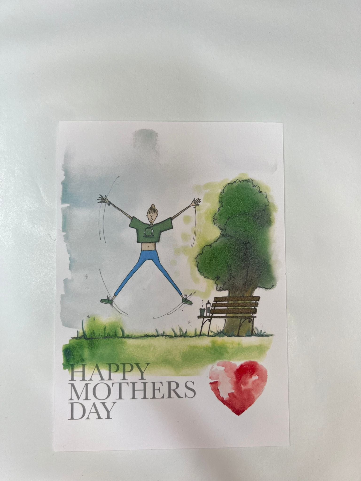 Blåbär Nordic Living’s Mothers Day Card - Fitness Mum features a joyful figure jumping near a park bench and tree in watercolor style. "HAPPY MOTHERS DAY" and a red heart add a warm, handmade touch to this unique Mother's Day card.