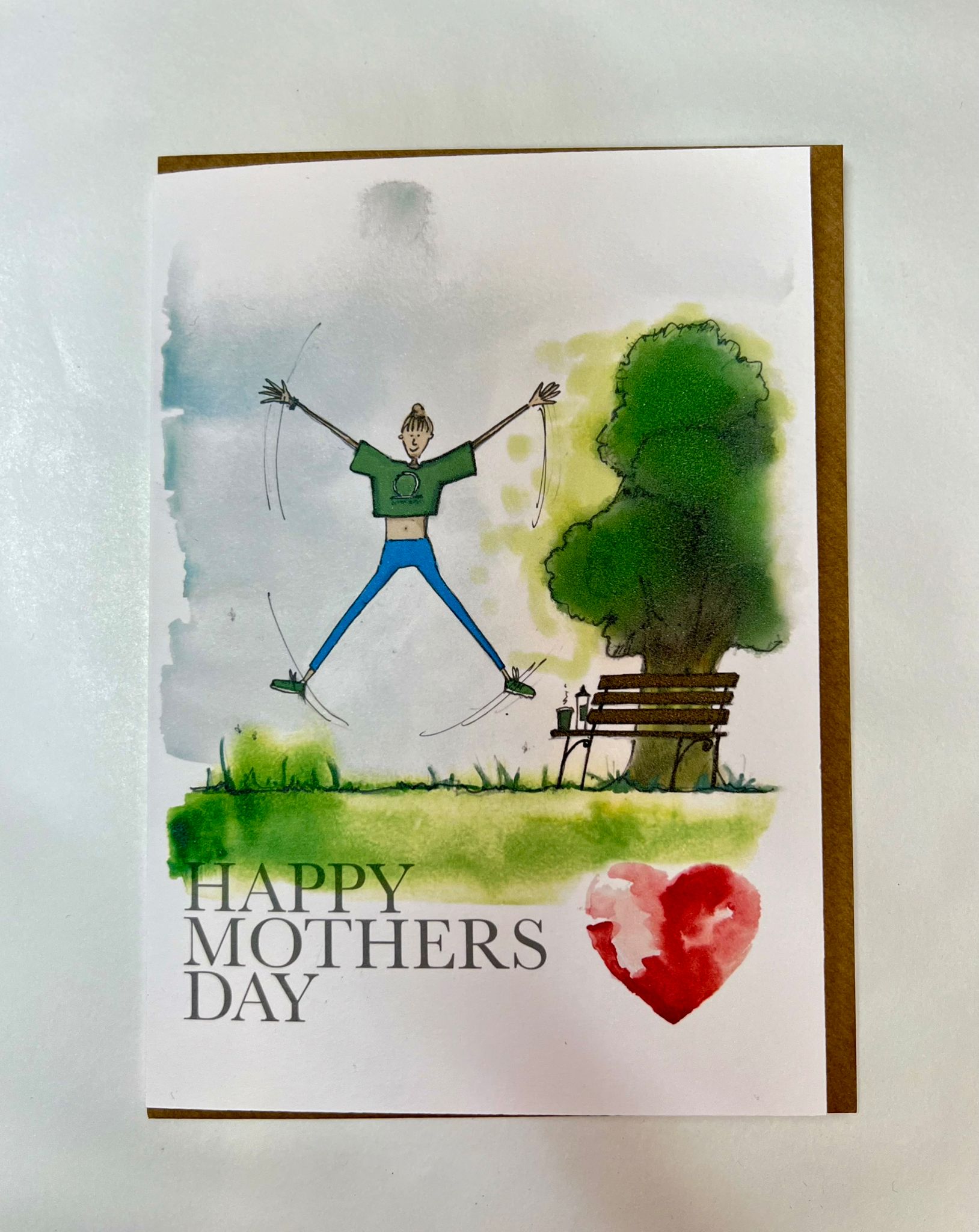 The Mothers Day Card - Fitness Mum by Blåbär Nordic Living features a handmade watercolor of a joyful person jumping by a park bench and tree, inspired by Nordic culture, with "HAPPY MOTHERS DAY" and a red heart painted at the bottom.