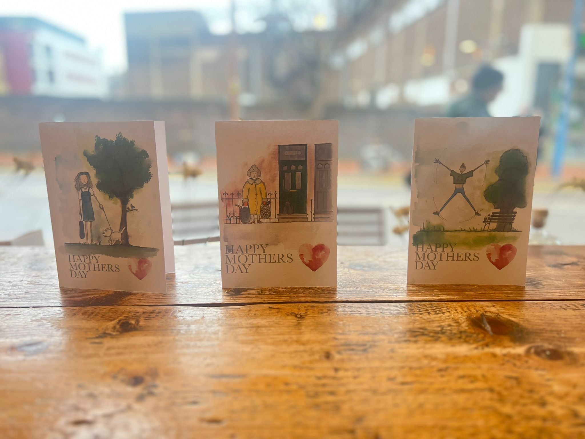 The Mothers Day Card - Fitness Mum by Blåbär Nordic Living stands upright on a wooden table near a window, featuring an illustrated heart and “Happy Mother’s Day” text, with a blurred outdoor backdrop.