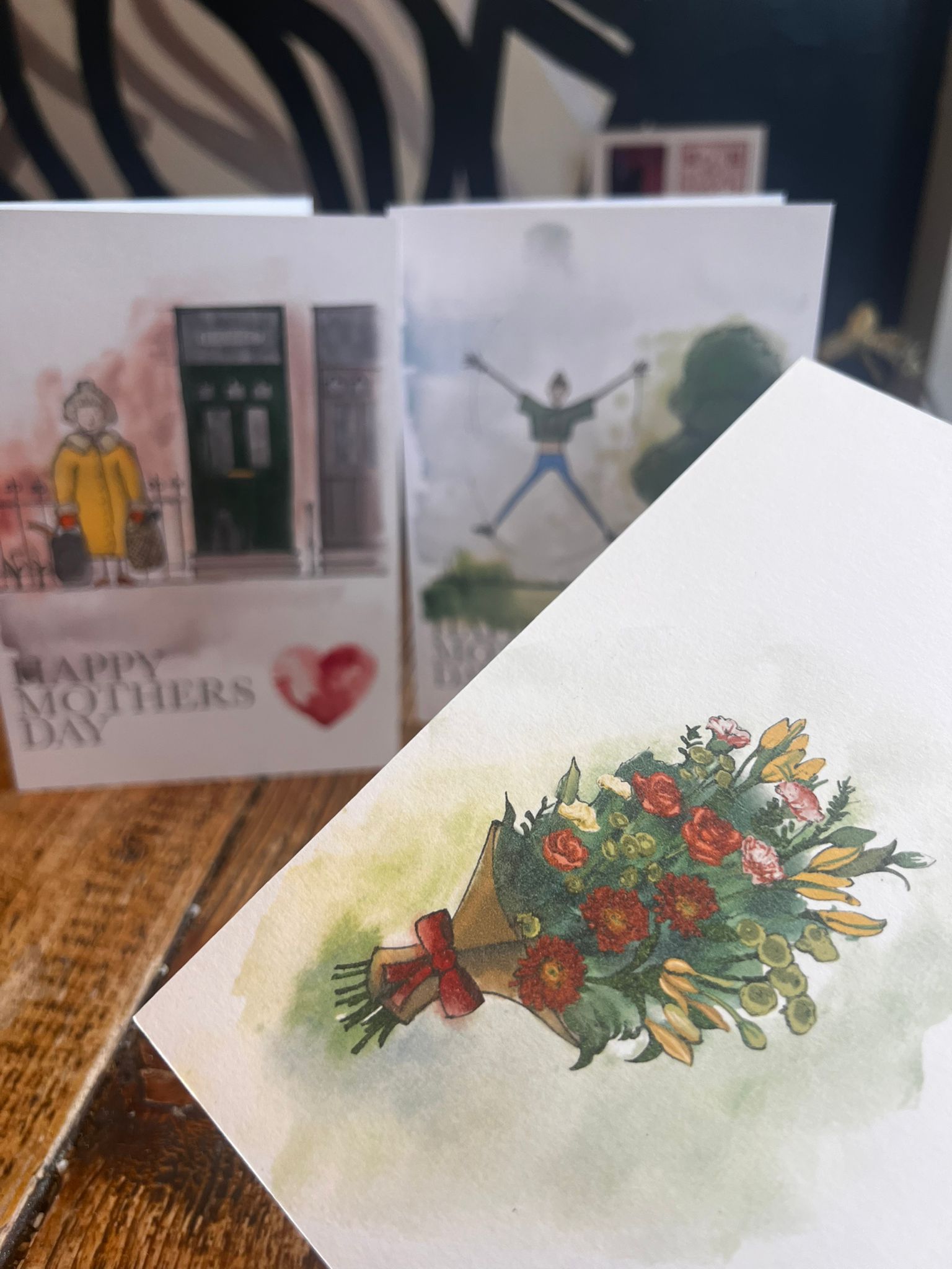 A close-up of the Blåbär Nordic Living "Mothers Day Card - Fitness Mum" on a wooden surface, with three other illustrated cards—one reading "Happy Mothers Day"—slightly blurred in the background.