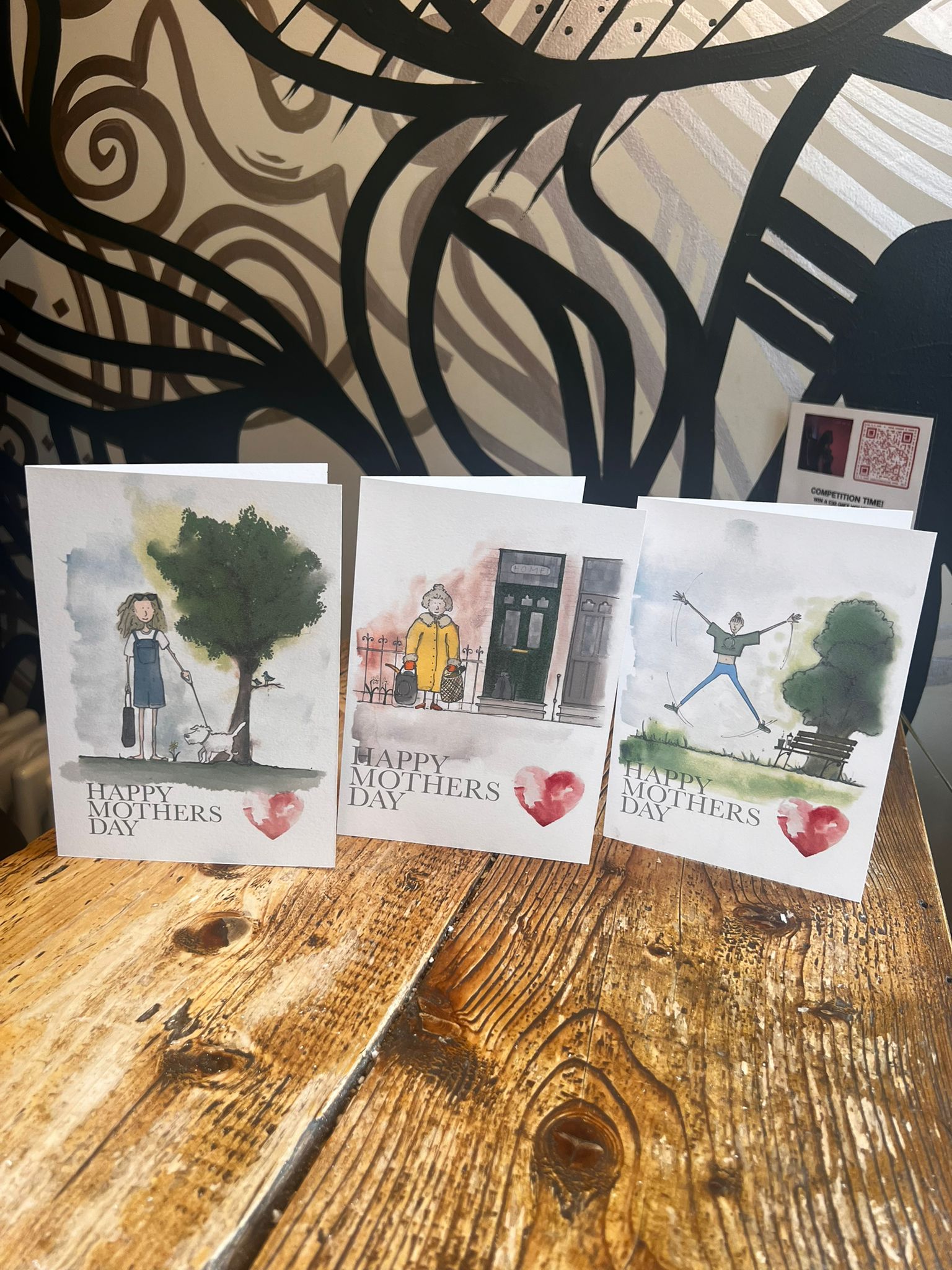 Three illustrated Mother’s Day cards from Blåbär Nordic Living, including the “Fitness Mum” design, stand on a wooden table. Each handmade card features an outdoor scene with women and “Happy Mothers Day” with a red heart on the front.