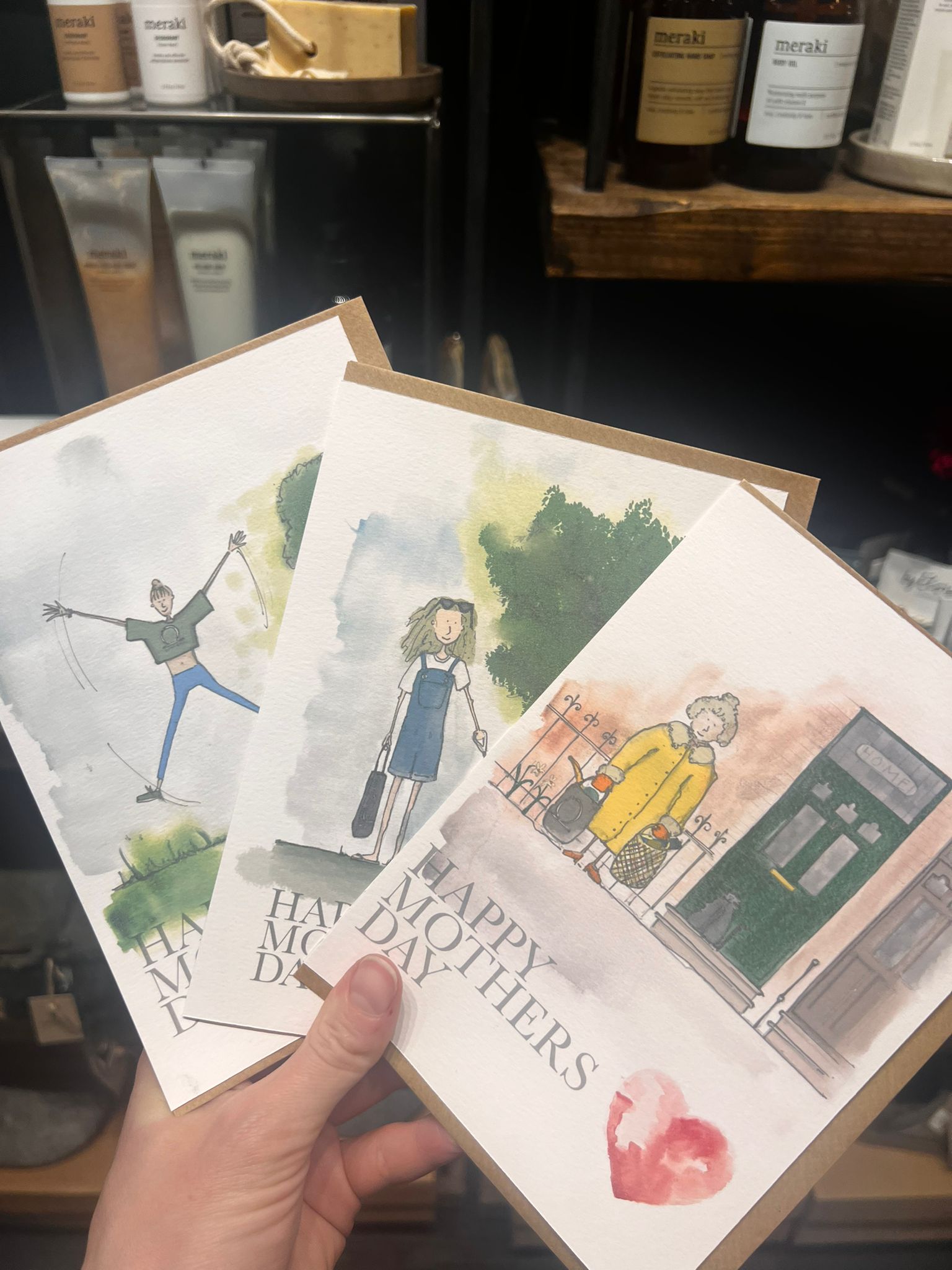 A hand holds three Blåbär Nordic Living "Mothers Day Card - Shopping Mum" cards, each showcasing unique watercolor art of women outdoors, with shelves of products blurred in the background.