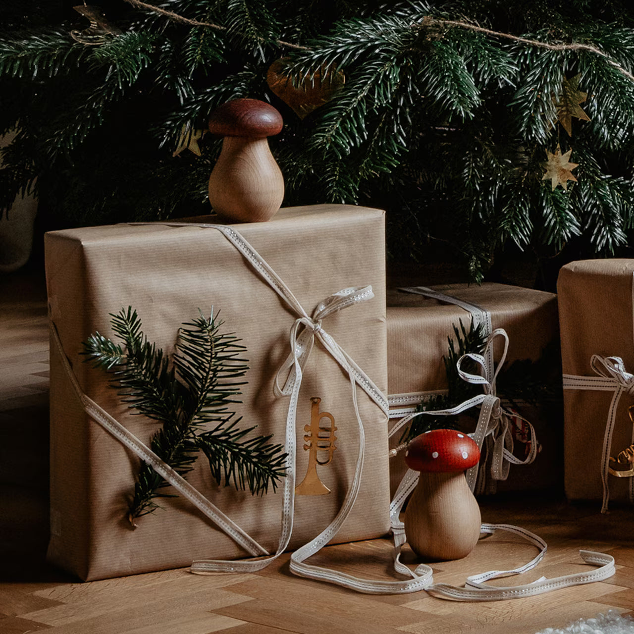 Brown paper-wrapped gifts with white ribbon and pine sprigs, featuring the Strömshaga Nutcracker Mushroom Fly Agaric (Flugsvamp) in beech wood, are placed under a Christmas tree.