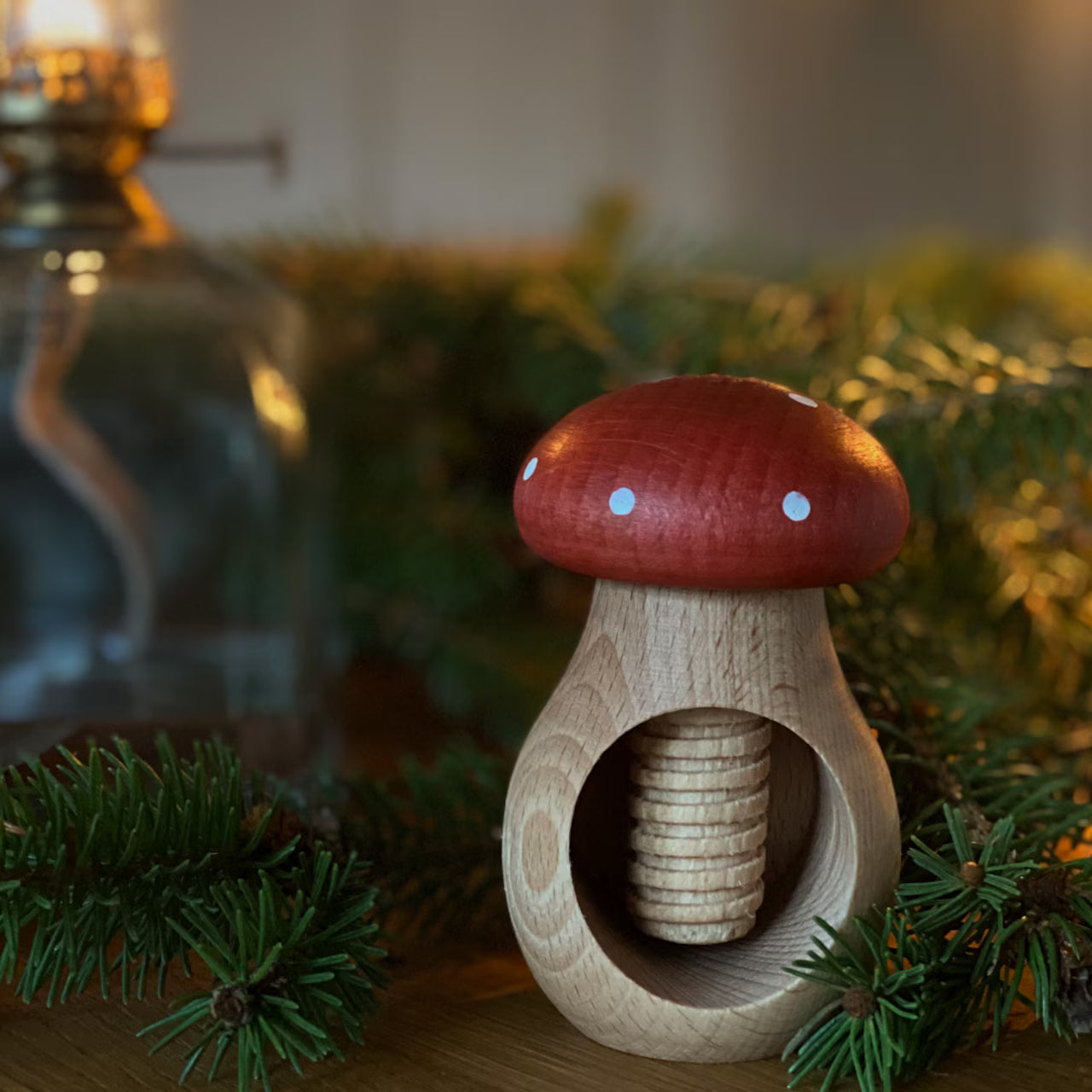 The Strömshaga Nutcracker Mushroom Fly Agaric, Flugsvamp in beech wood, rests on a table amid pine branches, a glass lamp in the background.