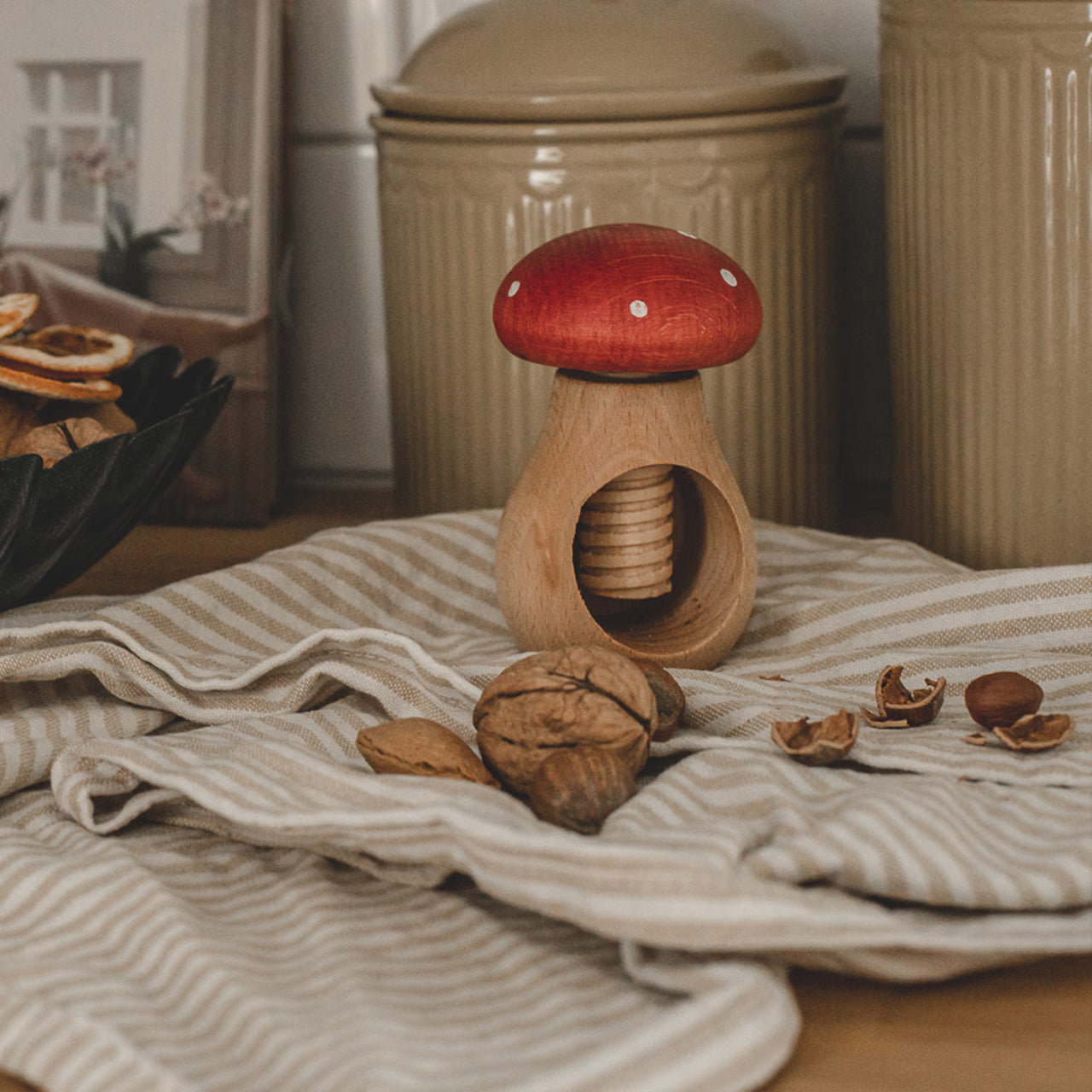 The Strömshaga Nutcracker Mushroom Fly Agaric (Flugsvamp) in beech wood, with a red cap, rests on a striped cloth with whole and cracked walnuts nearby, set in a cozy kitchen with jars and dried orange slices in the background.