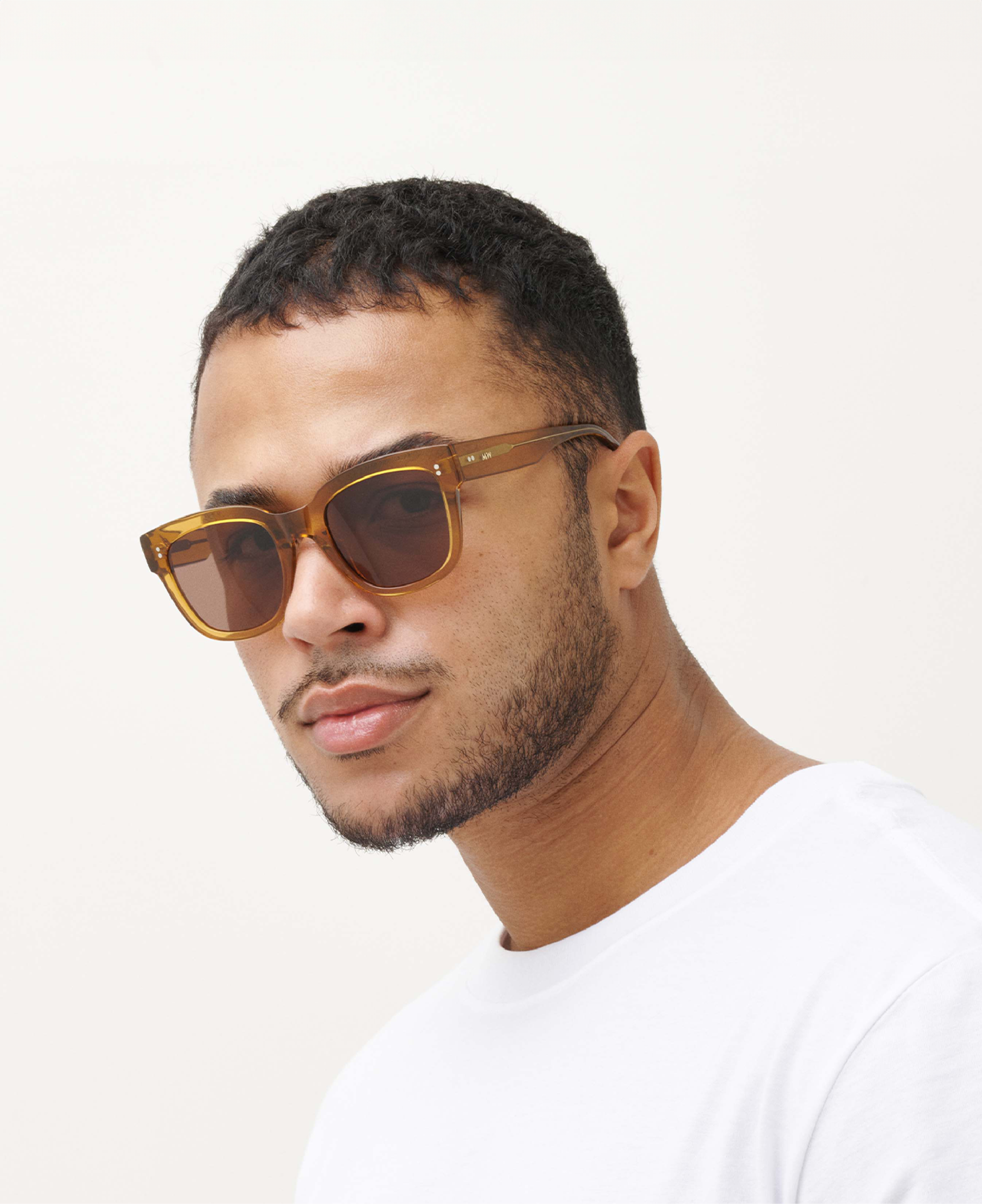 A man with short dark hair and facial hair wears MESSYWEEKEND Sunglasses Liv in Coffee Brown with brown lenses and a white t-shirt against a plain light background.