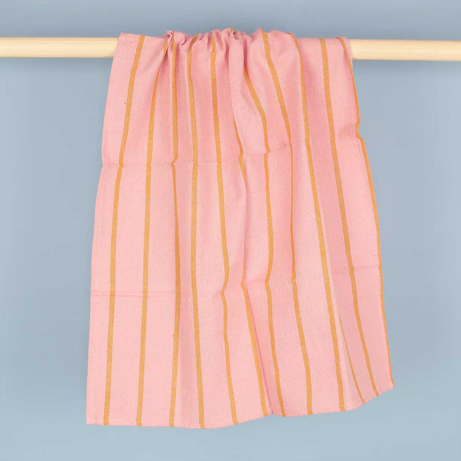 The Tea Towel, Wide Stripe Pink & Mustard in organic cotton by A World of Craft is handwoven and draped over a light wooden rod, displayed against a plain light blue background.