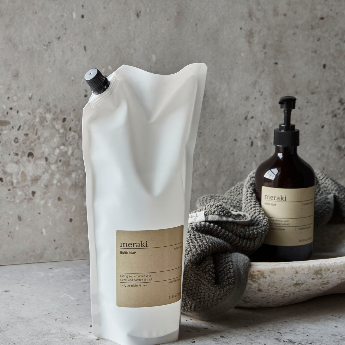 A Meraki Hand Soap Organic, Northern Dawn Refill pouch stands upright next to a Meraki soap pump bottle and a folded towel in a ceramic dish on a stone surface.