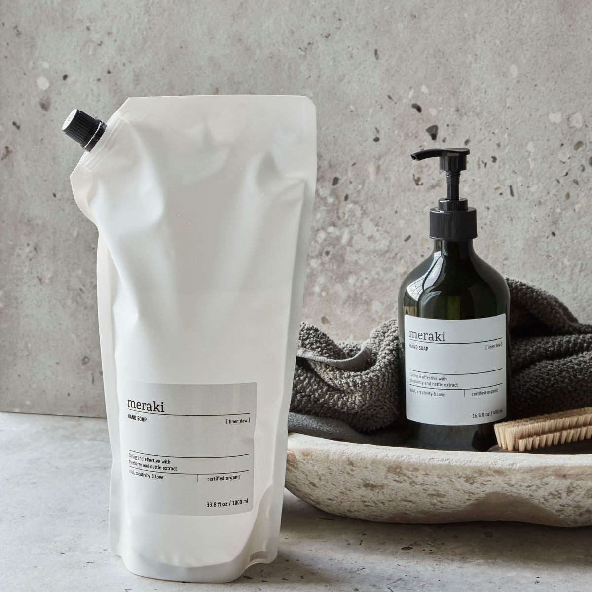 A Meraki Hand Soap Organic, Linen Dew Refill pouch and a dark green Meraki bottle sit on a stone surface, with a textured bowl, towel, and brush in the background.