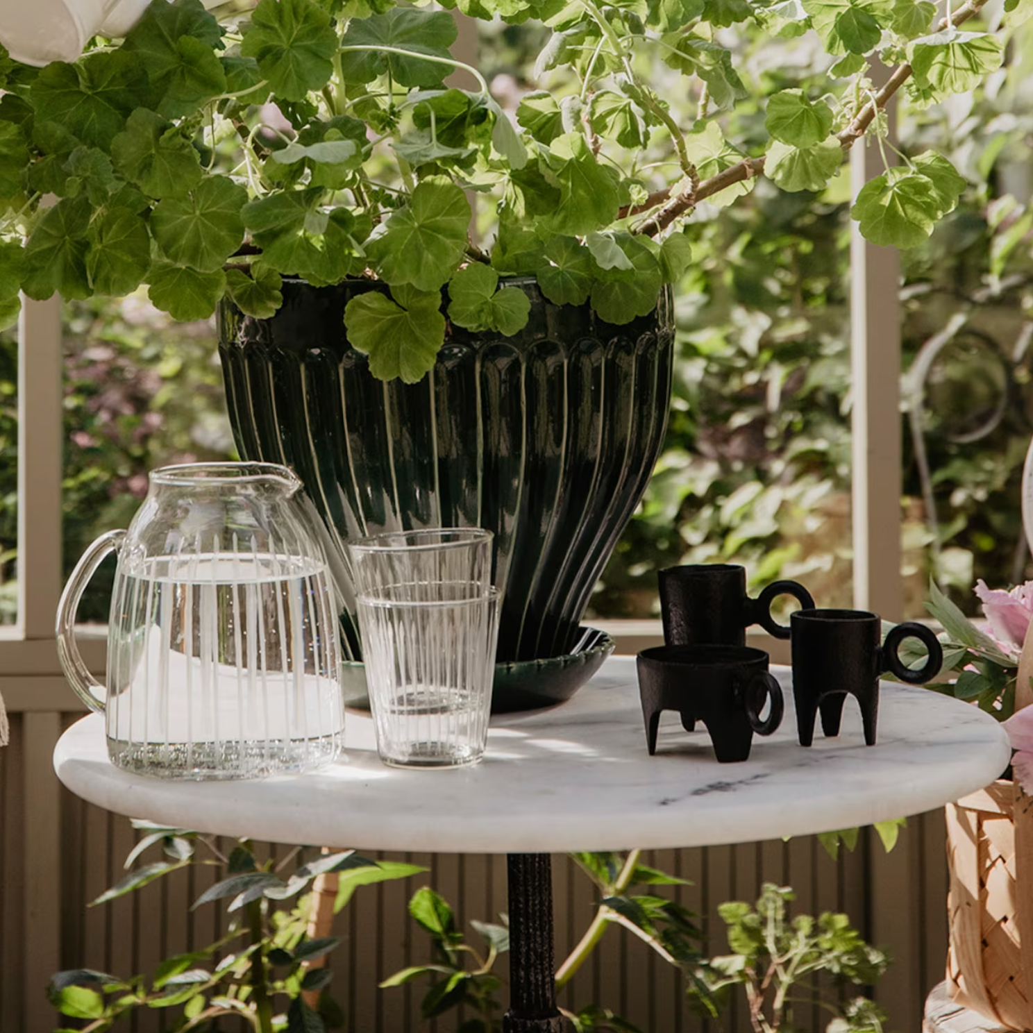 A round white table in a sunlit garden holds a glass pitcher of water, a drinking glass, three small black cups, Strömshagas cast iron Tea Light Holder Three Feet for large tea lights (6cm x13cm), and a large green potted plant.