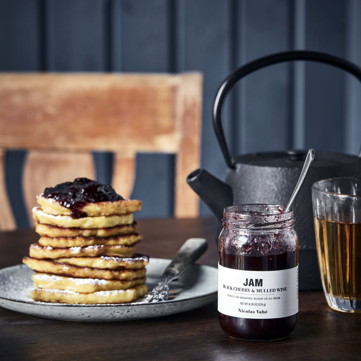 A stack of pancakes topped with Nicolas Vahé Jam Seasonal - Black Cherry & Mulled Wine sits on a plate beside the jar, accompanied by a glass of tea and a black teapot on a rustic wooden table.