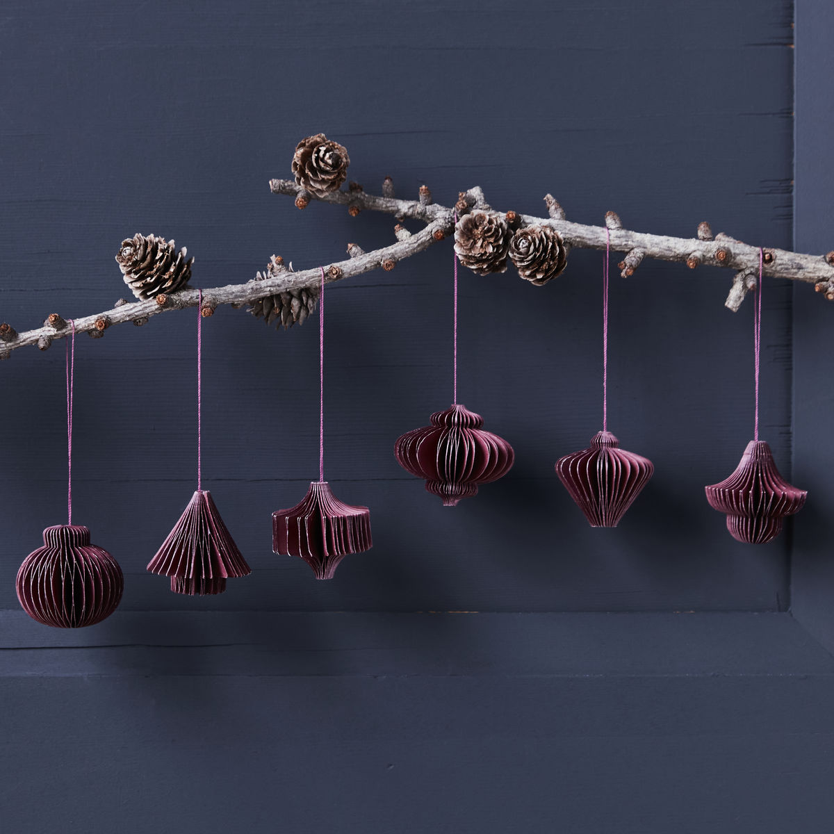 A branch with pinecones and the House Doctor Ornament Comb in Paper, Burgundy (set of 6) hangs against a dark blue wall, creating elegant Christmas décor.