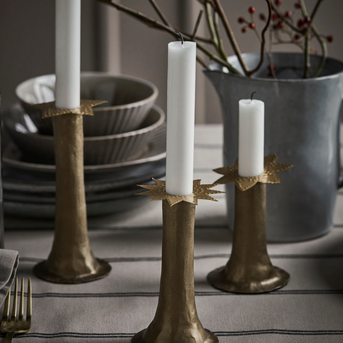 The Candle Rings Star in Brass, Set of 3 by House Doctor are displayed on a striped tablecloth beside stacked dishes and a metal jug with branches, bringing festive Christmas charm with their star-shaped bases.