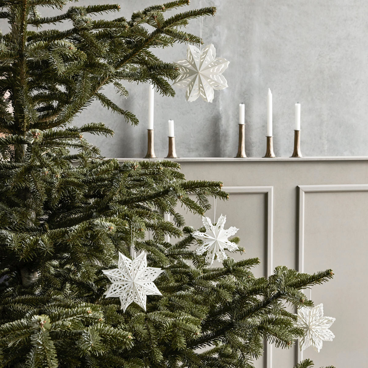 A Christmas tree with House Doctor’s Paper Snowflake Ornaments (set of 4) stands by a mantel with five white candles in metallic holders, creating modern holiday décor.