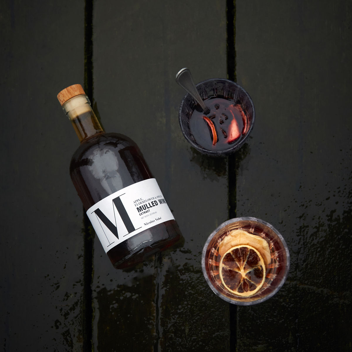 A bottle of Nicolas Vahé Mulled Wine Extract – Apple, Elderflower & Spices sits on a dark wet wooden surface beside two festive Christmas drink glasses, one with a spoon and the other topped with citrus slices.
