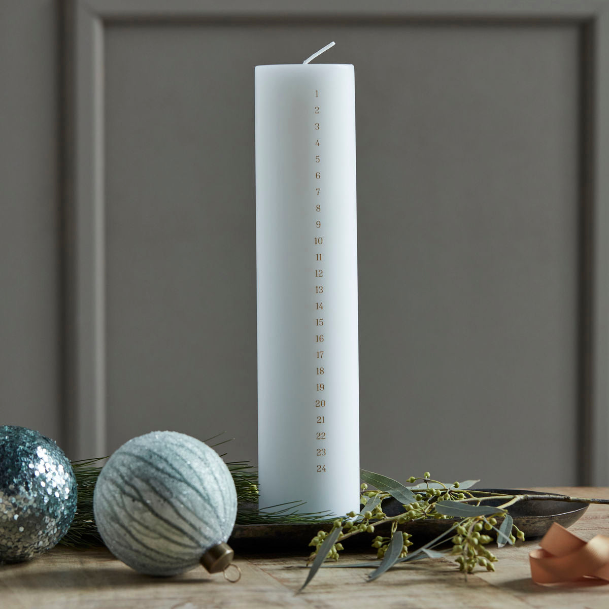 The House Doctor Calendar Candle 24 Days of Christmas, Forest in White (6cm), stands upright on a wooden surface, numbered 1 to 24 for a festive countdown amid ornaments, greenery, and ribbon.
