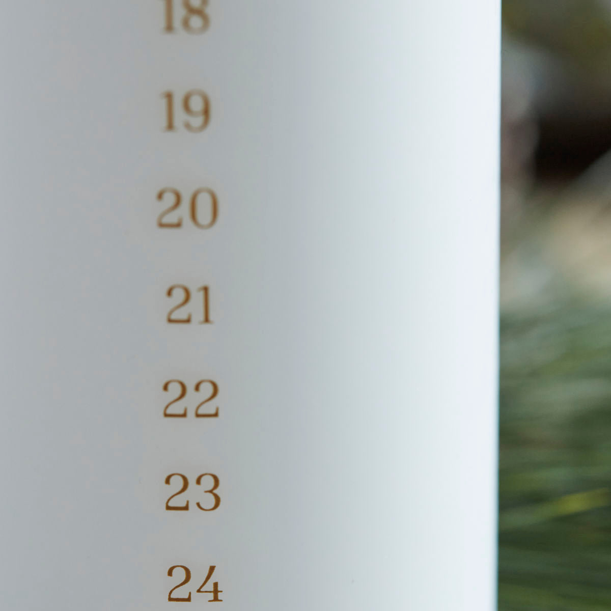 A close-up of the House Doctor Calendar Candle 24 days of Christmas, Forest in White (6cm), shows gold numbers 18–24 for a festive countdown. Blurred greenery accents the background.