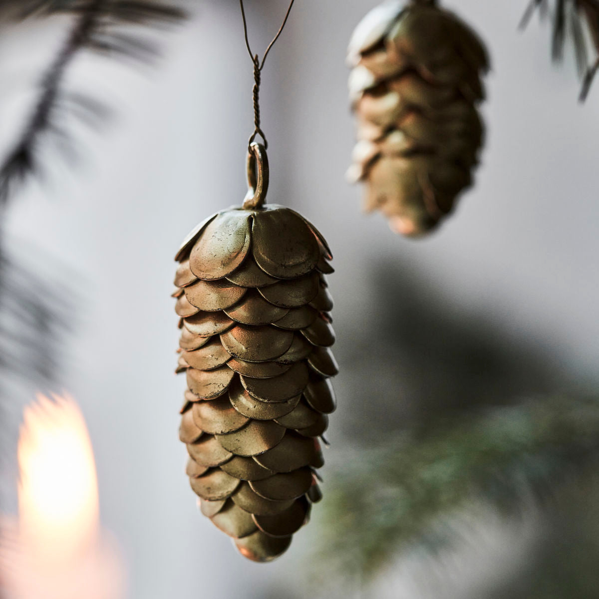 A close-up of House Doctor's Ornaments Pinecones in Iron, Set of 2, displays elegant metallic pine cone décor on a branch, with the second ornament softly blurred in the background.