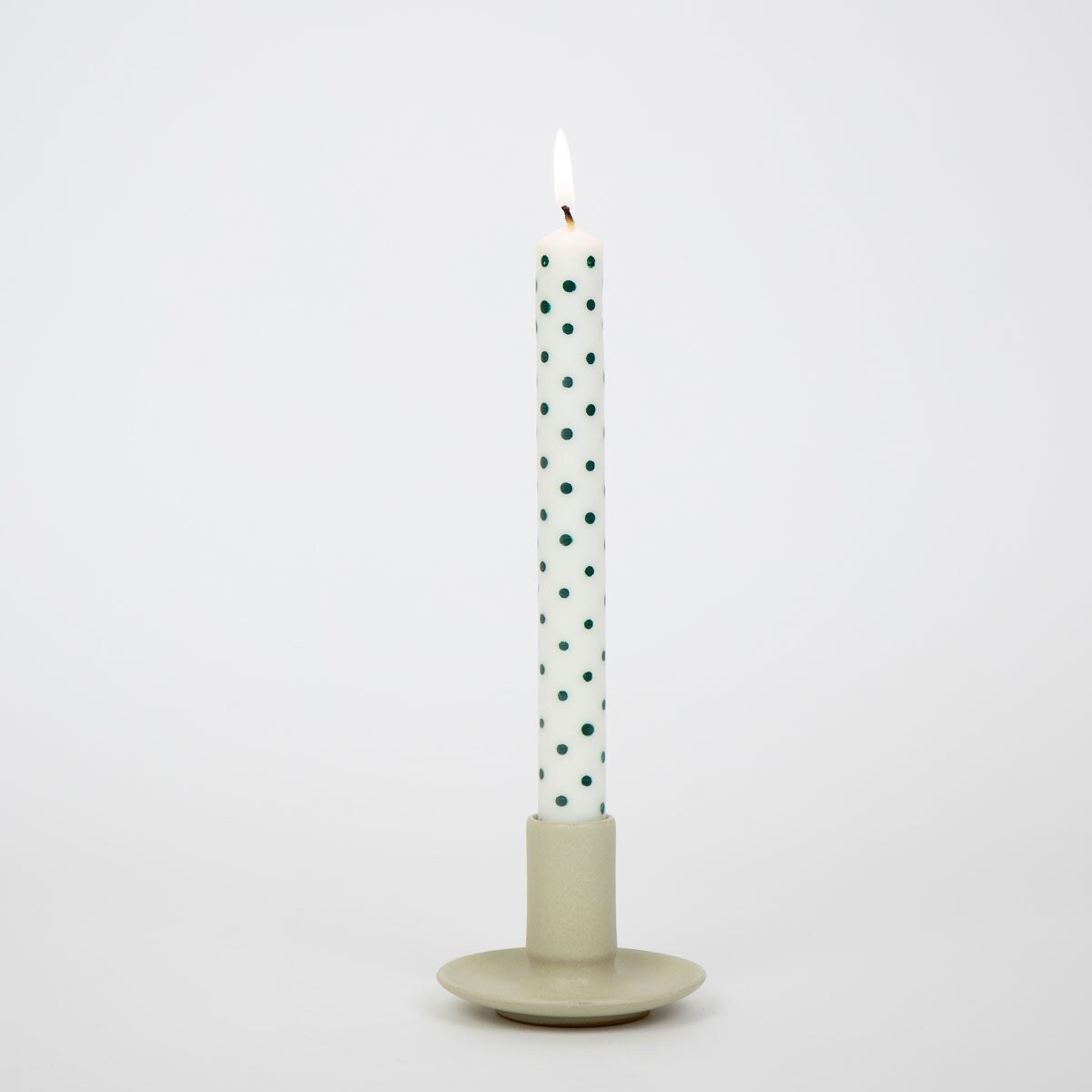 A lit, hand-painted Candles Minidot White & Green, Handmade 2 Pack taper candle with small green polka dots in a simple ceramic candle holder.