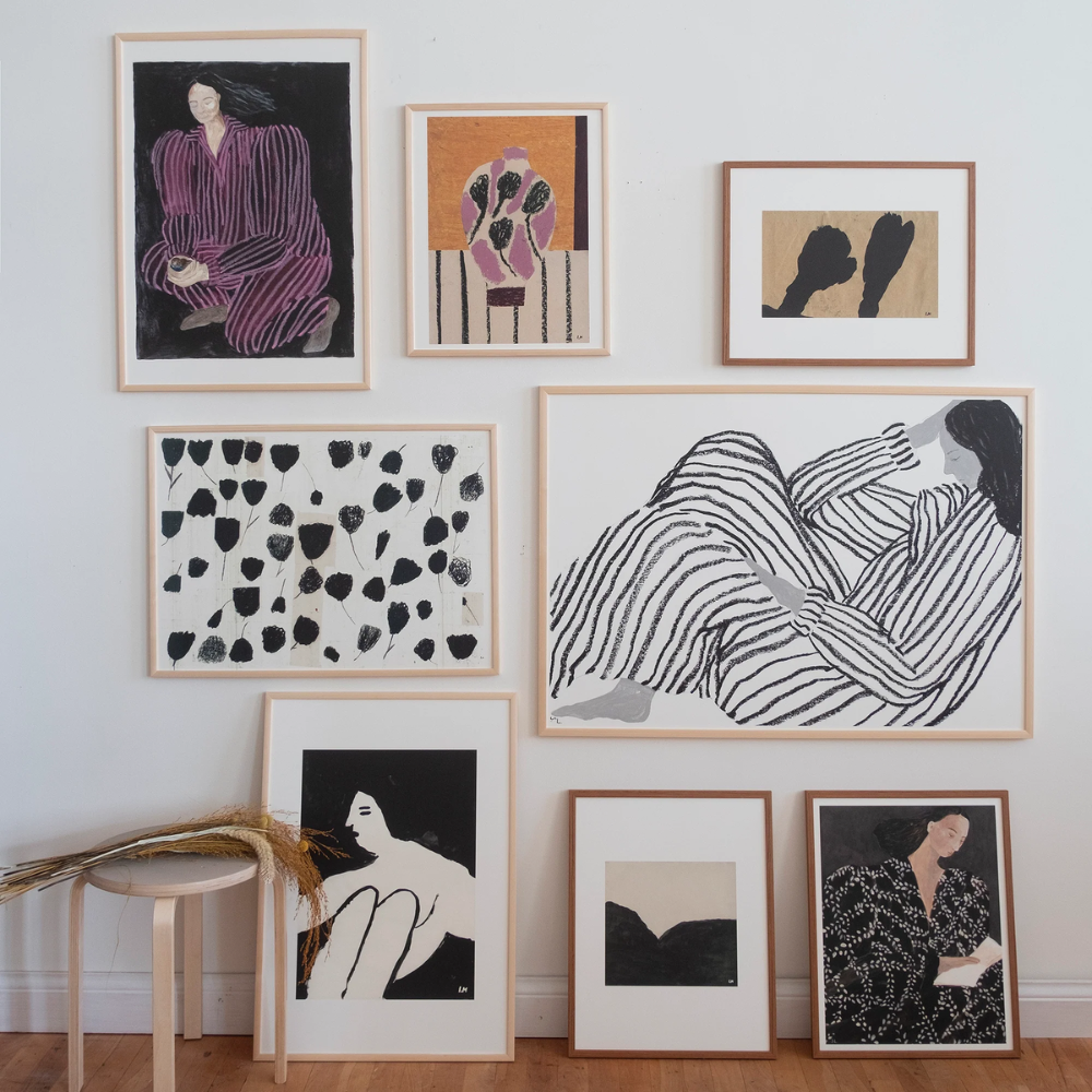A gallery wall displays eight framed art prints featuring abstract and minimalist illustrations of human figures, mostly women, in black, white, and muted earthy tones. A stool with dried grass is placed below the arrangement.