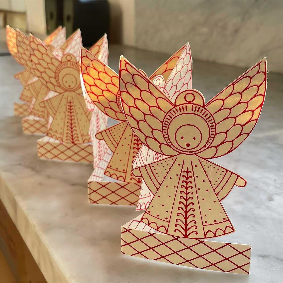 Four Folklore Angel Paper Foldout Christmas Decorations by A World of Craft stand in a row on a marble surface, their red line designs glowing warmly from behind—a charming handmade Scandinavian accent perfect as a festive angel display or tabletop decoration.