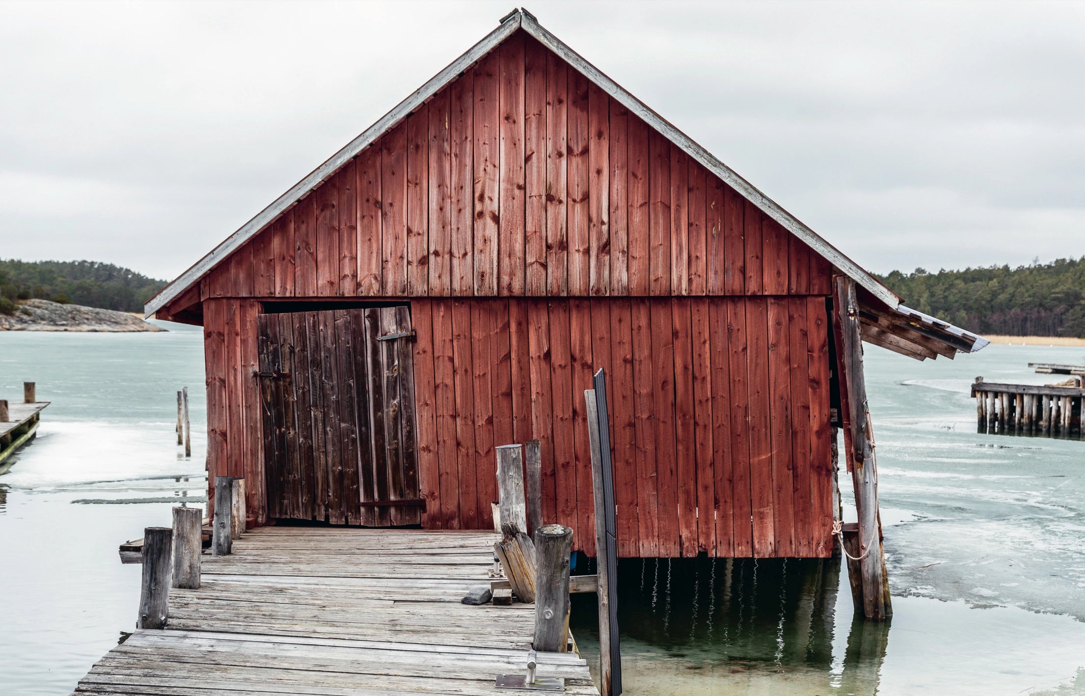 The book "Genuine – Handmade Beauty from the North" by Cozy Publishing features a weathered red boathouse on stilts over ice, connected by a crooked dock, with an overcast sky and forested land in the background.