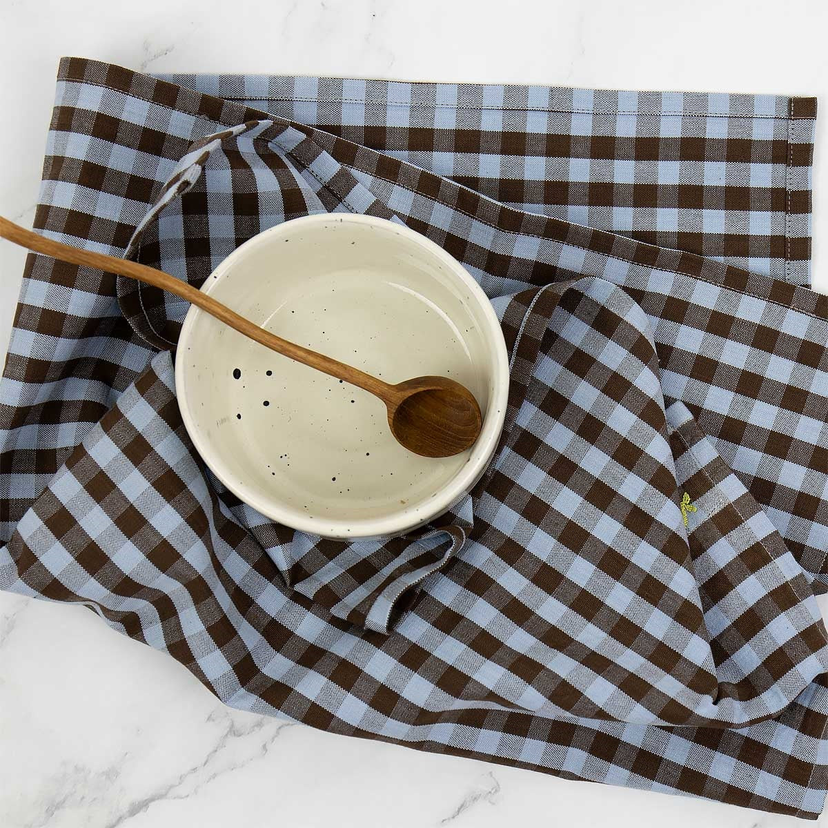 A ceramic bowl with a wooden spoon sits on the Maria Towel, Light Blue & Brown in Organic Cotton by A World of Craft, displayed on a white marble surface.