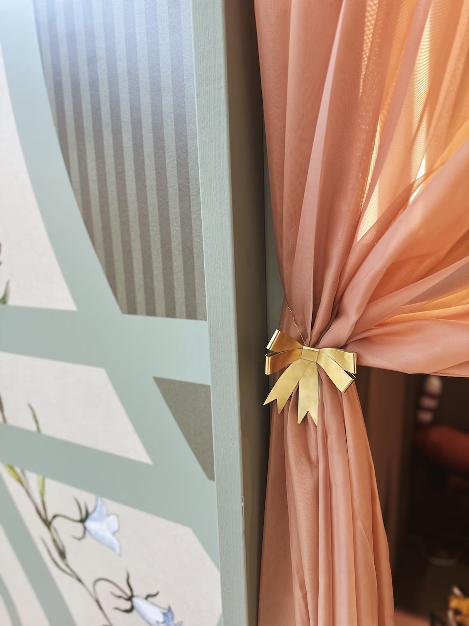 A peach-colored sheer curtain is tied with the Strömshaga Ribbon Hanging Ornament in brass beside a decorative wall panel featuring green geometric stripes and painted white flowers.