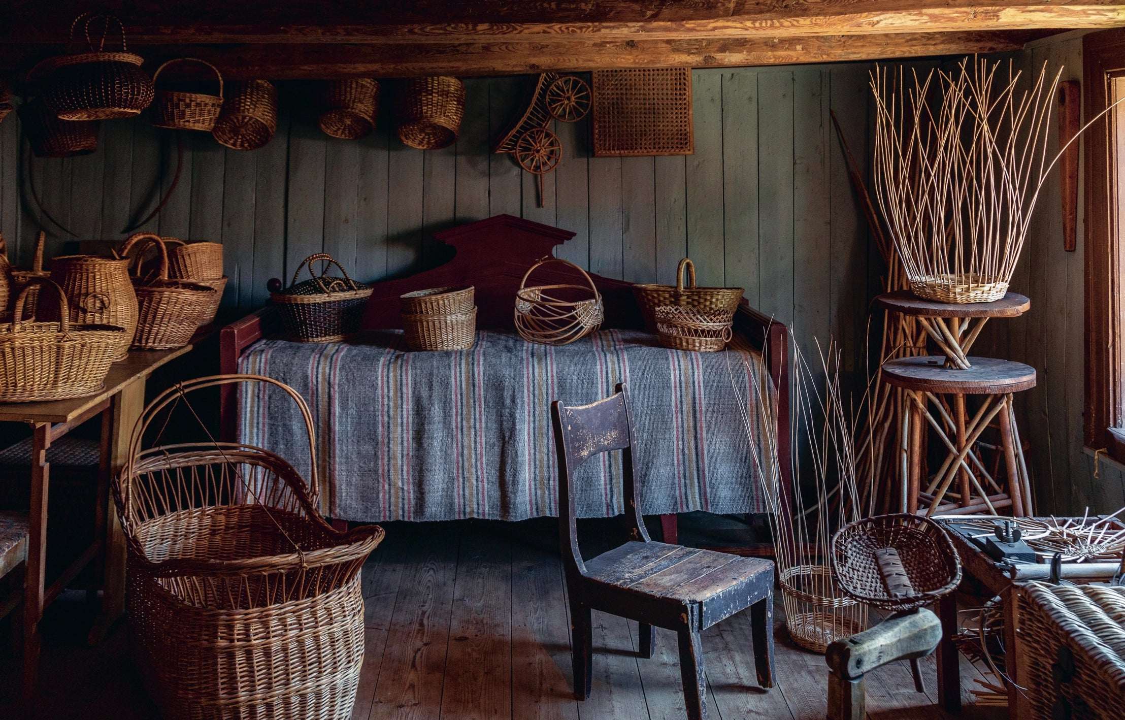 A rustic room with wooden walls and wicker baskets reflects Finnish artisanal heritage, as featured in "Genuine – Handmade Beauty from the North" by Cozy Publishing. A striped bed cover and chair bask in natural light from the window.