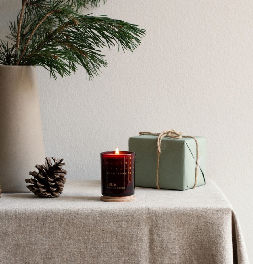 A pinecone, a Skandinavisk Seasonal Jul Scented Candle 65g in a glass holder, and a gift wrapped with twine sit on a beige cloth-covered table; in the background is a vase filled with pine branches.