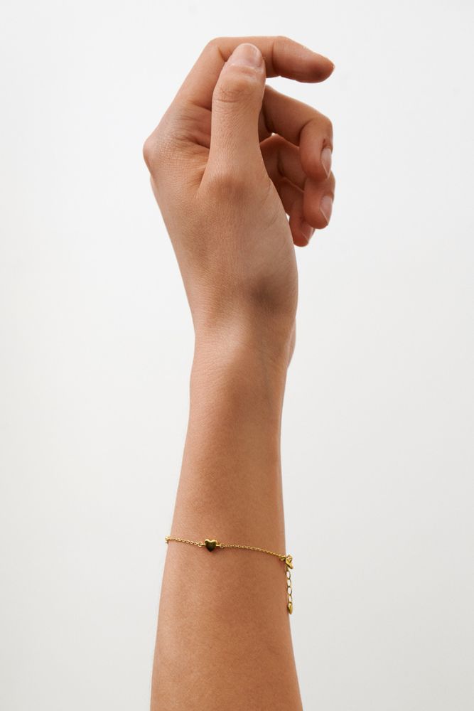 A hand with slightly curled fingers is raised against a plain white background, wearing the Pernille Corydon Adore Bracelet in Gold—a minimalistic gold chain bracelet with a delicate heart charm.