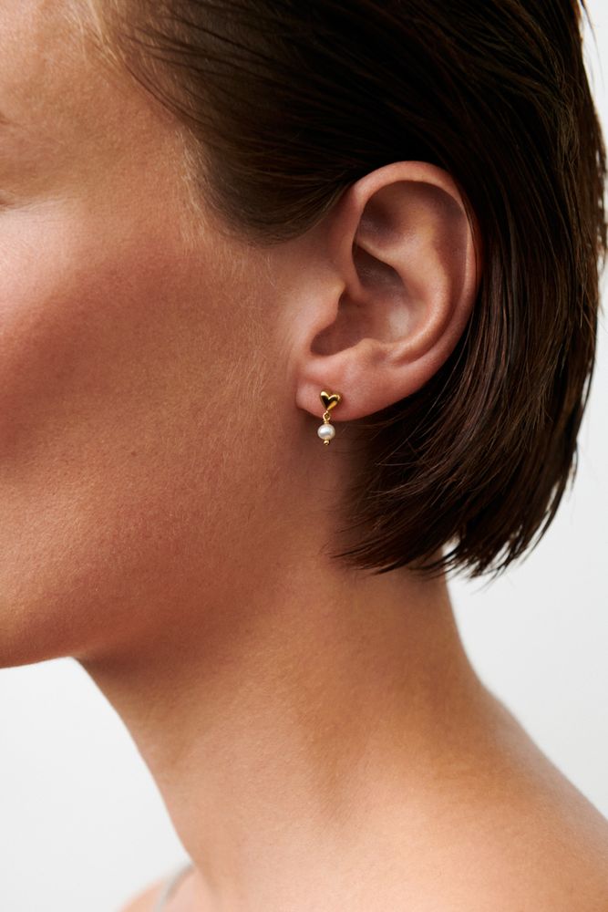 Close-up of a person with short brown hair wearing Pernille Corydon's White Adore Earrings in Gold, each featuring a heart stud and a dangling freshwater pearl, set against a plain, light background.