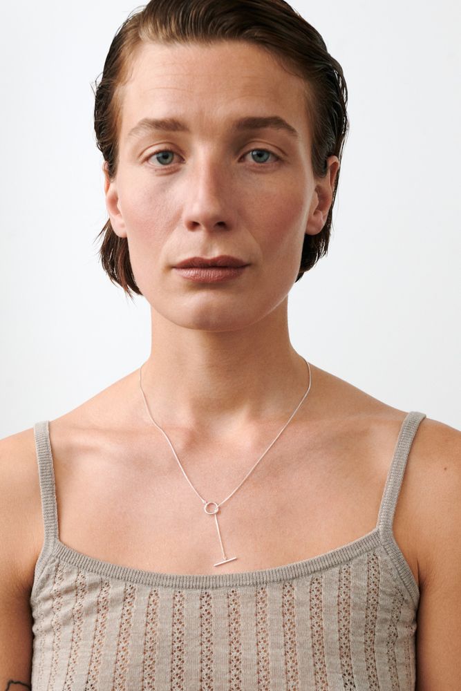 A person with short brown hair and blue eyes wears a beige textured tank top and the Pernille Corydon Hope Necklace in Gold, featuring a circular T-bar clasp, looking at the camera against a plain white background.