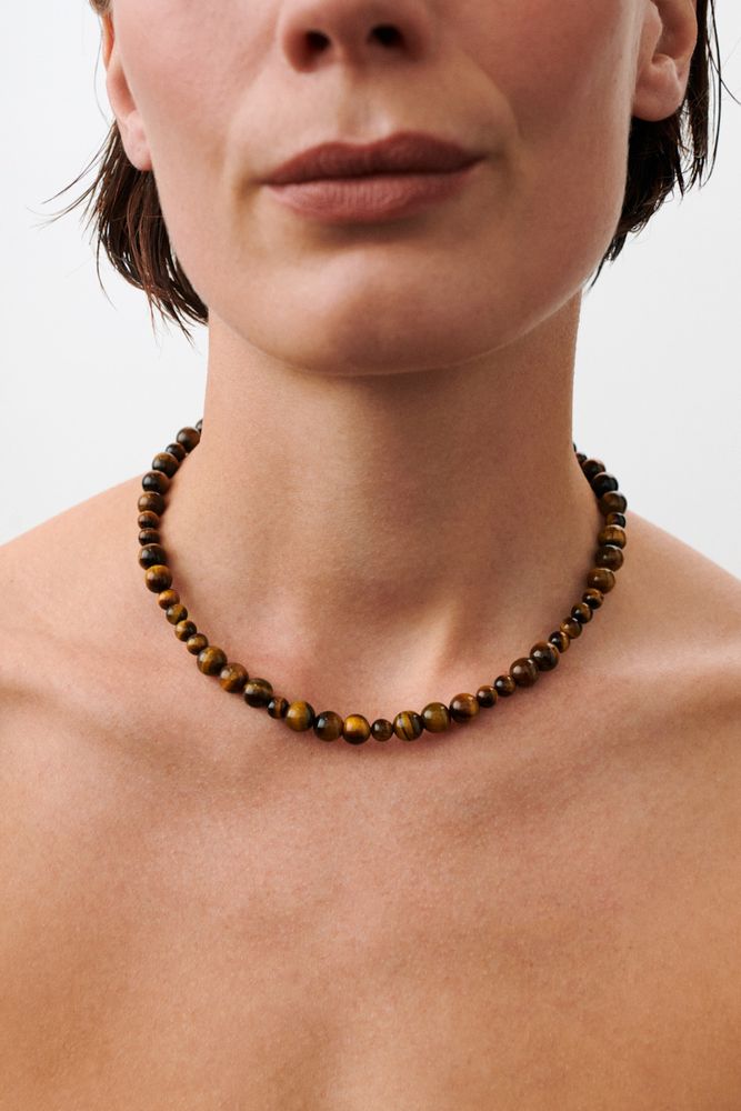 A close-up of a person wearing the Pernille Corydon Earth Necklace with Tiger Eye stones, showing the lower half of their face and neck. The necklace features beads in brown shades resembling natural Tiger Eye gems against a plain background.