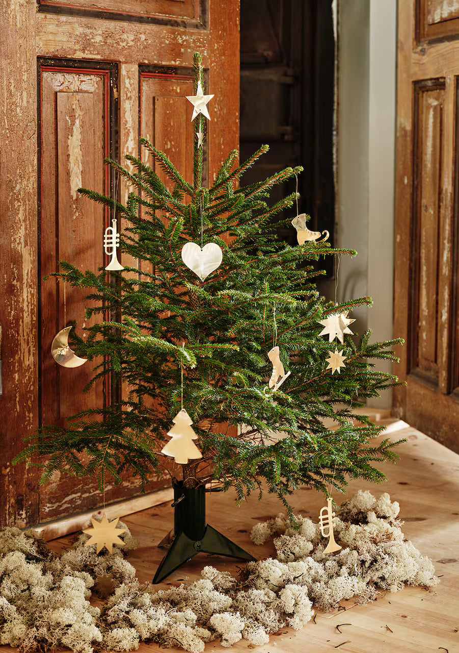 A small Christmas tree decorated with Strömshaga’s brass Hanging Ornaments (set of 6) stands on a wooden floor with white moss, positioned before rustic wooden doors.