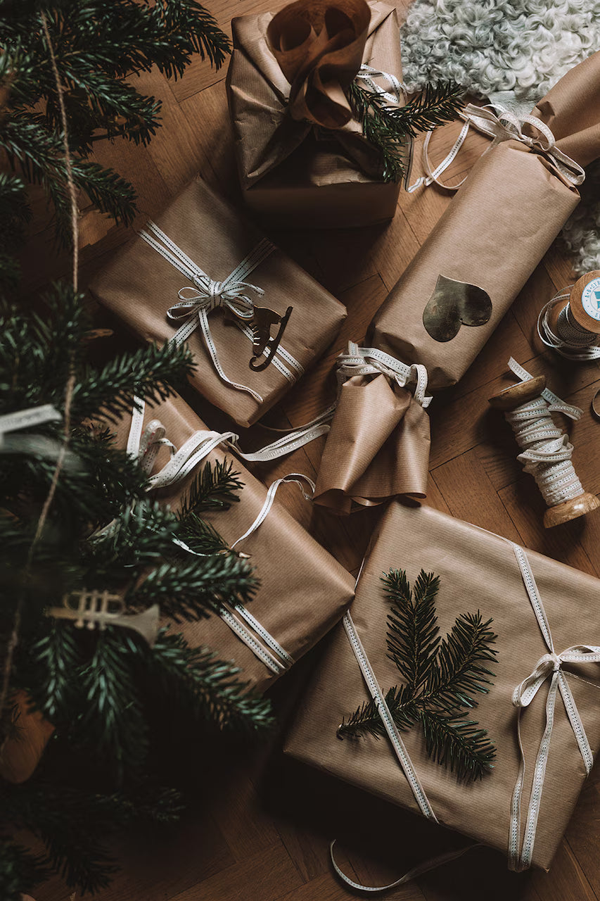 Hanging Ornaments, Set of 6, Christmas, in Brass by Strömshaga are displayed on a wooden floor with gifts wrapped in brown paper, white ribbon, evergreen branches, classic Christmas motifs, and a spool of string.